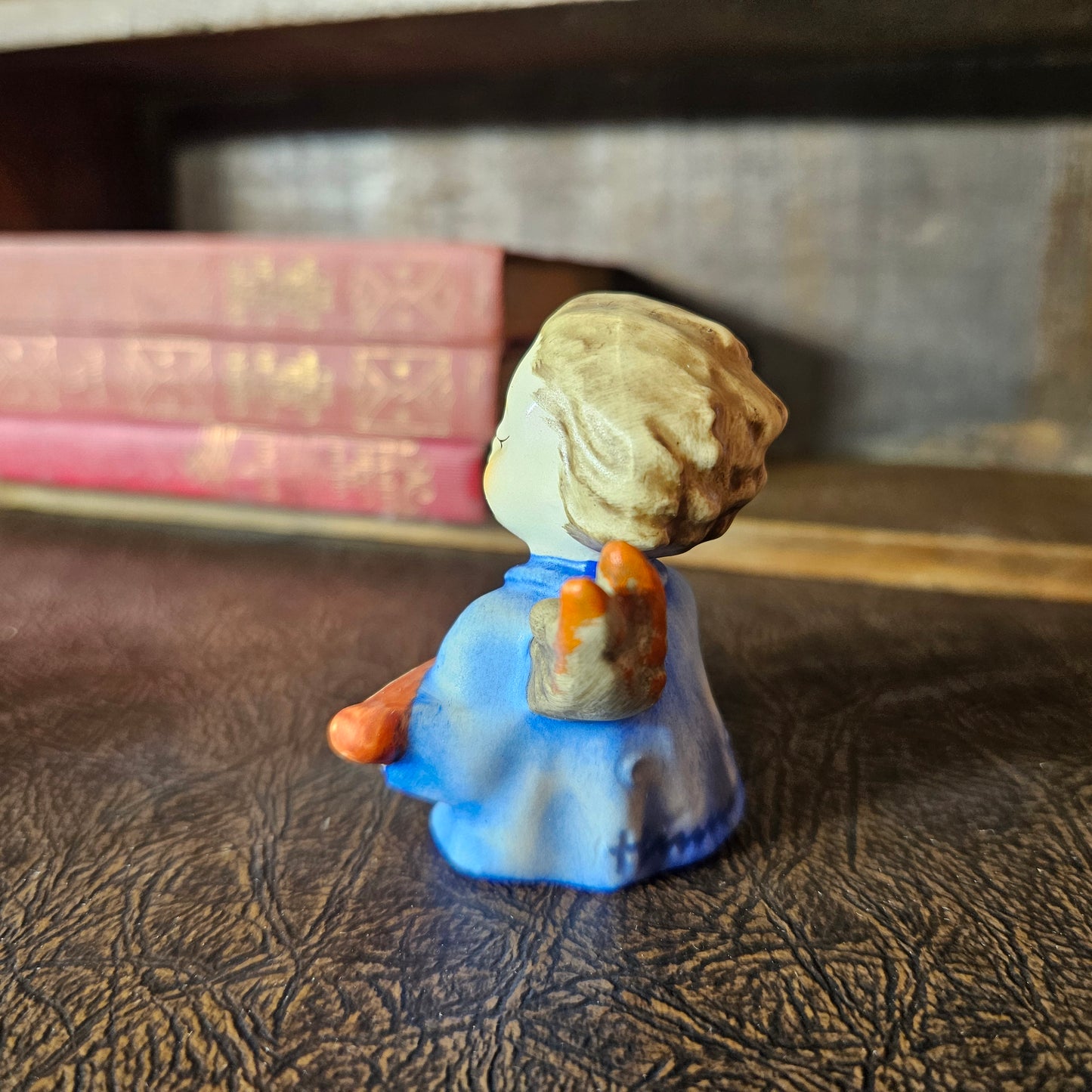 Small figurine of a angel in a blue dress with a brown bag on a textured surface with books in the background.