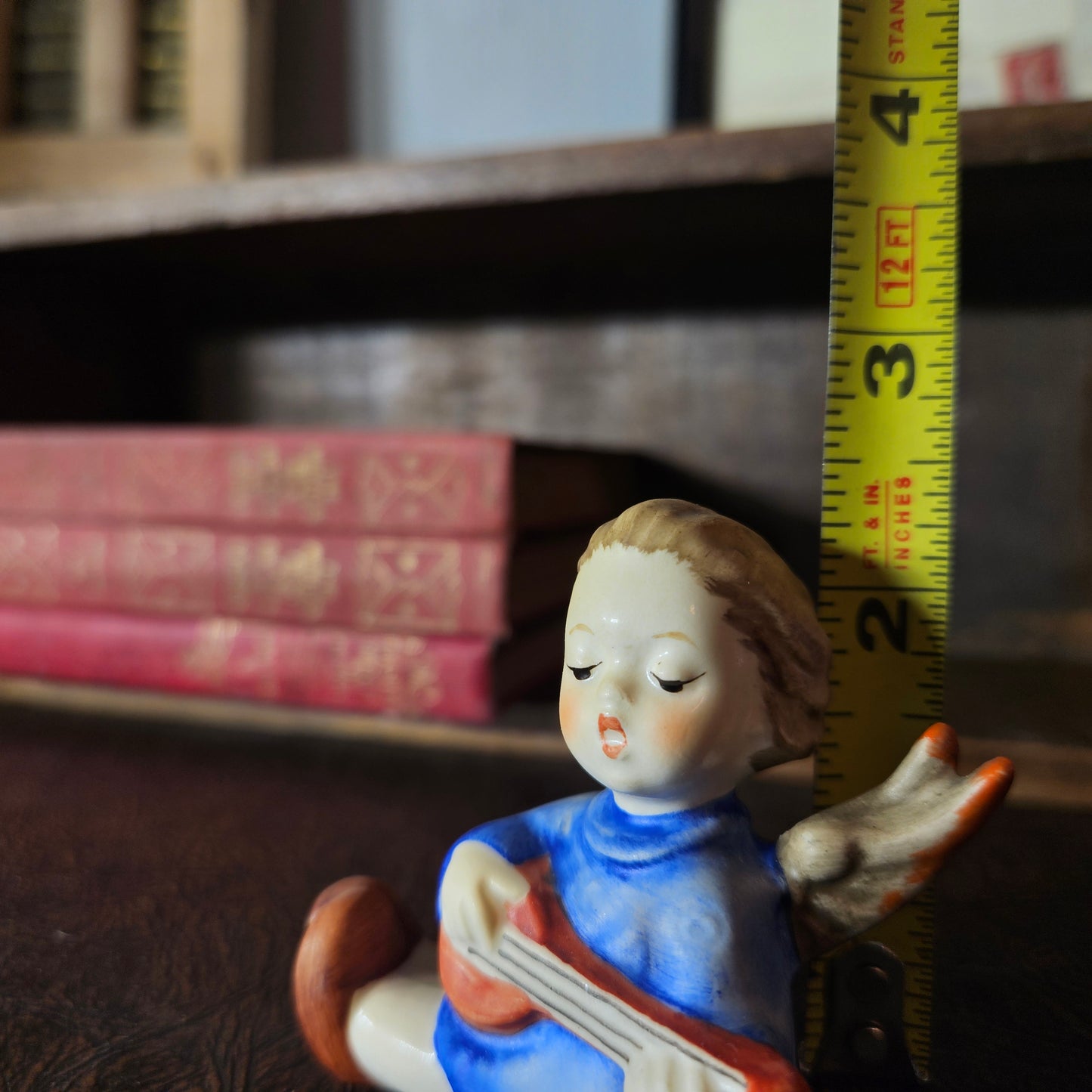 Vintage figurine of a girl playing a harp with a tape measure for scale on a bookshelf background.