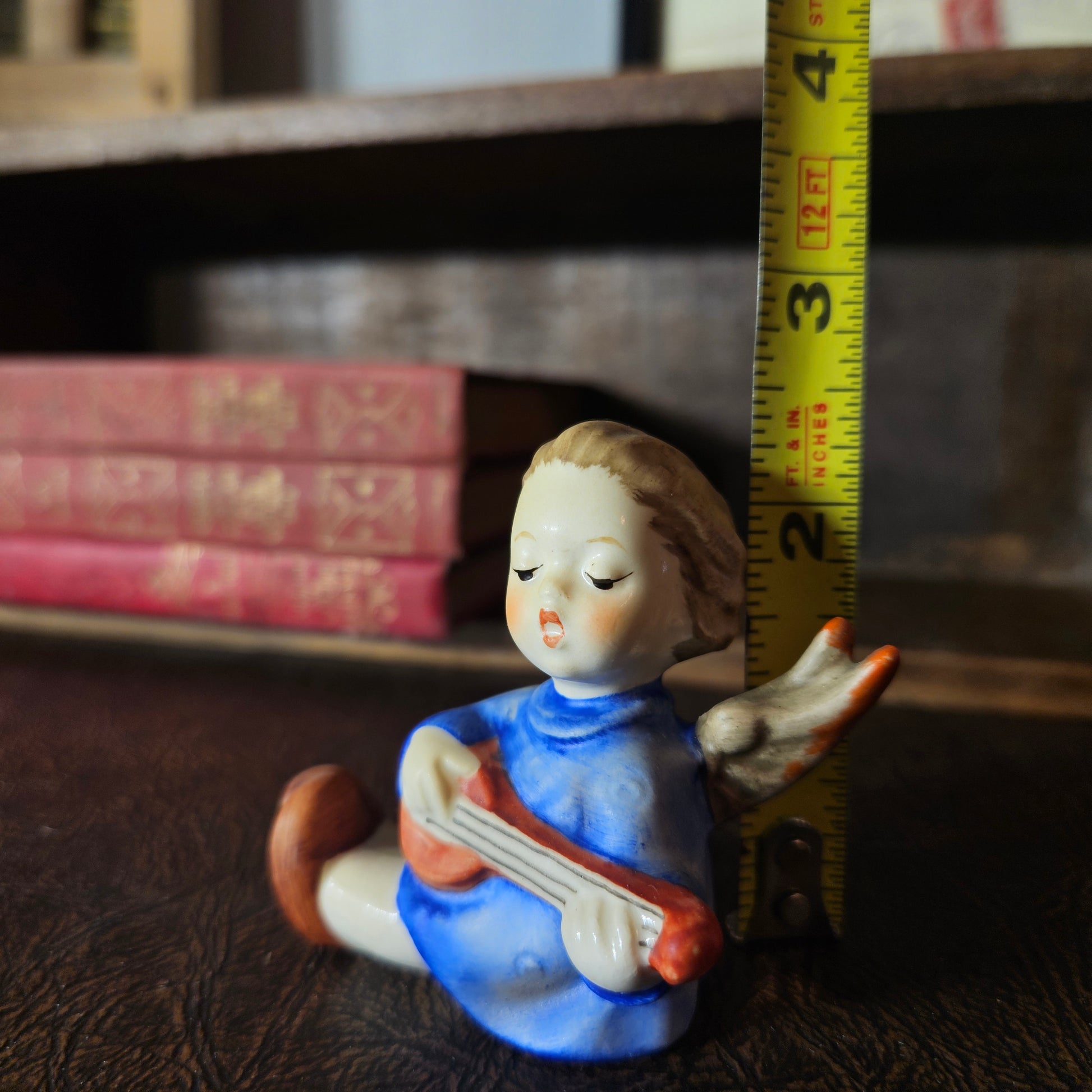Small figurine of a girl playing a lyre with a measuring tape for scale, on a wooden surface with books in the background.