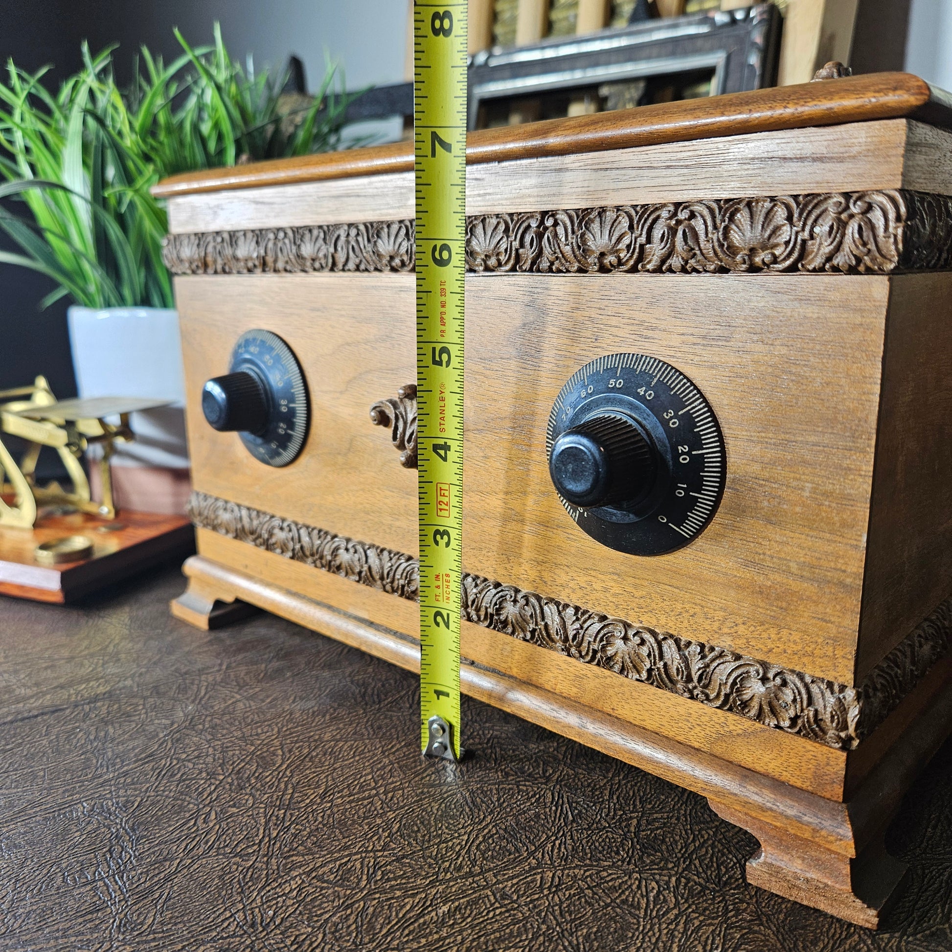 Wooden music box with decorative carvings and a tape measure for scale on a textured surface.