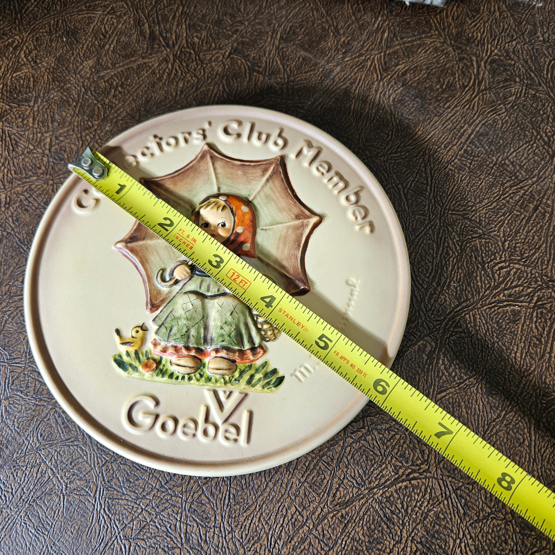 Decorative plate with Goebel branding and a measuring tape for scale on a textured surface