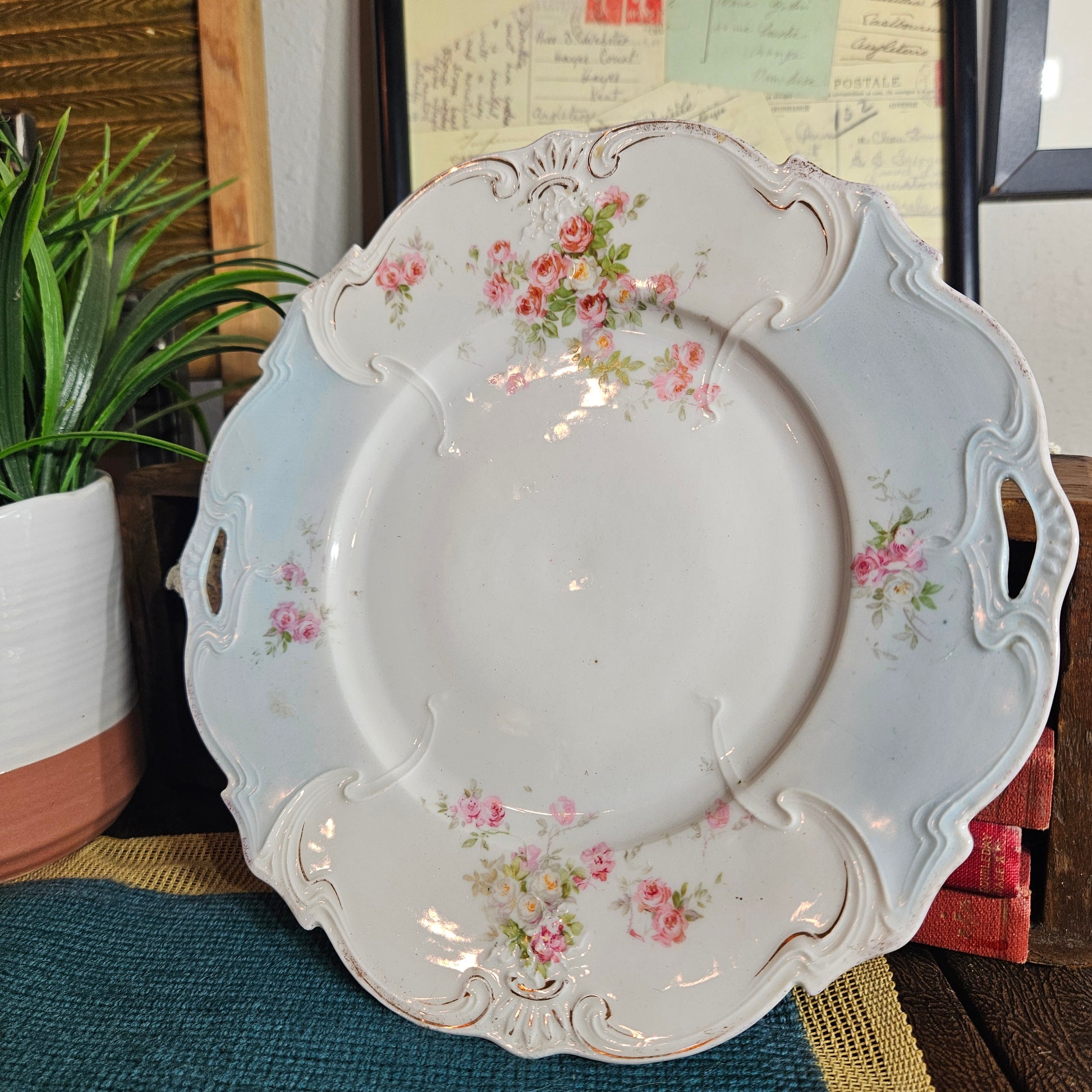 Decorative white tray with floral patterns on a surface with a plant and framed pictures in the background.