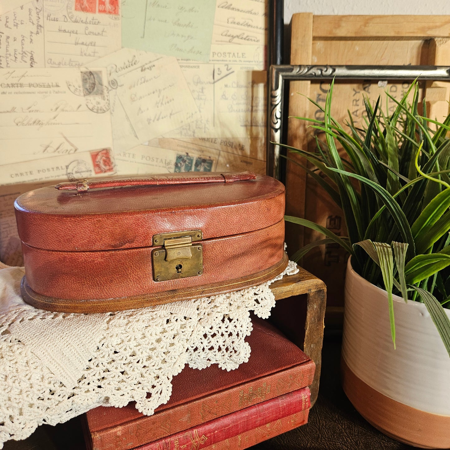 Vintage-style brown leather suitcase on a lace-covered surface with a plant and framed postcards in the background.