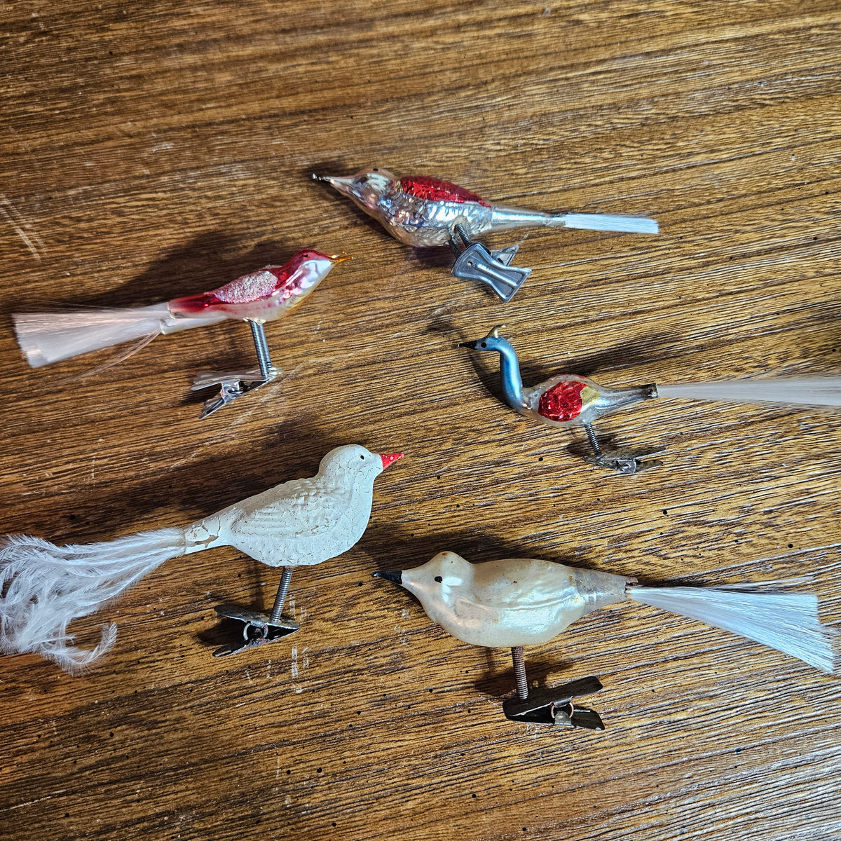 Five vintage bird-shaped pocket watches with metal spring-clip closures are arranged on a wooden tabletop. The birds feature white, red, blue, and cream-colored bodies with white feather details.