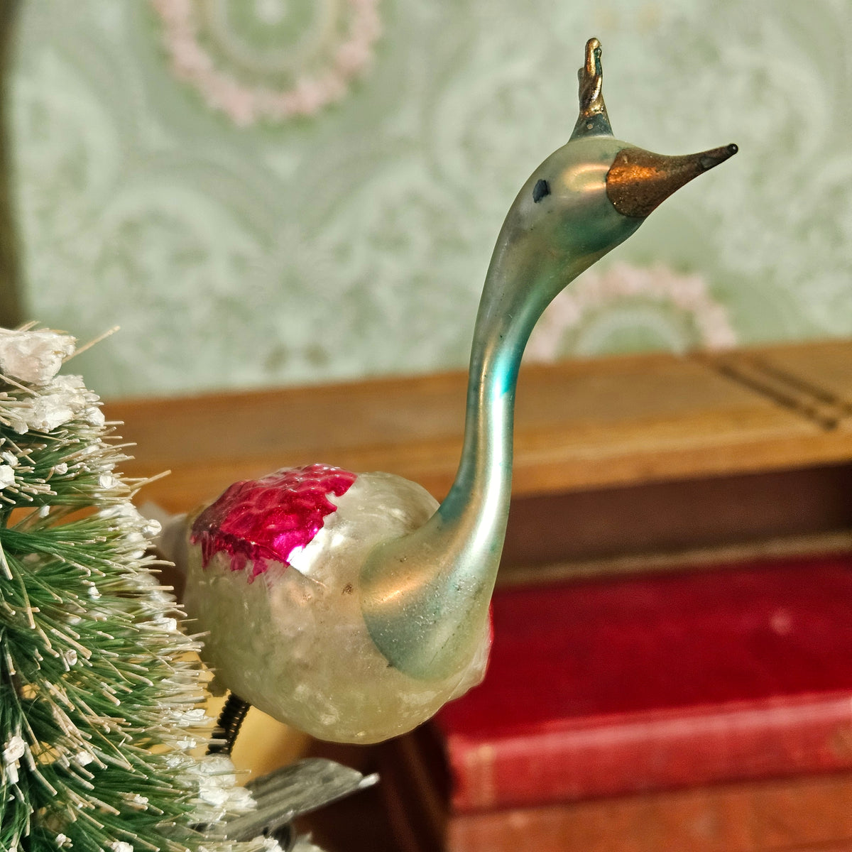 A vintage glass swan ornament with a metallic teal neck, copper beak, and a pink glittery chest patch, shown close-up next to a small artificial tree.