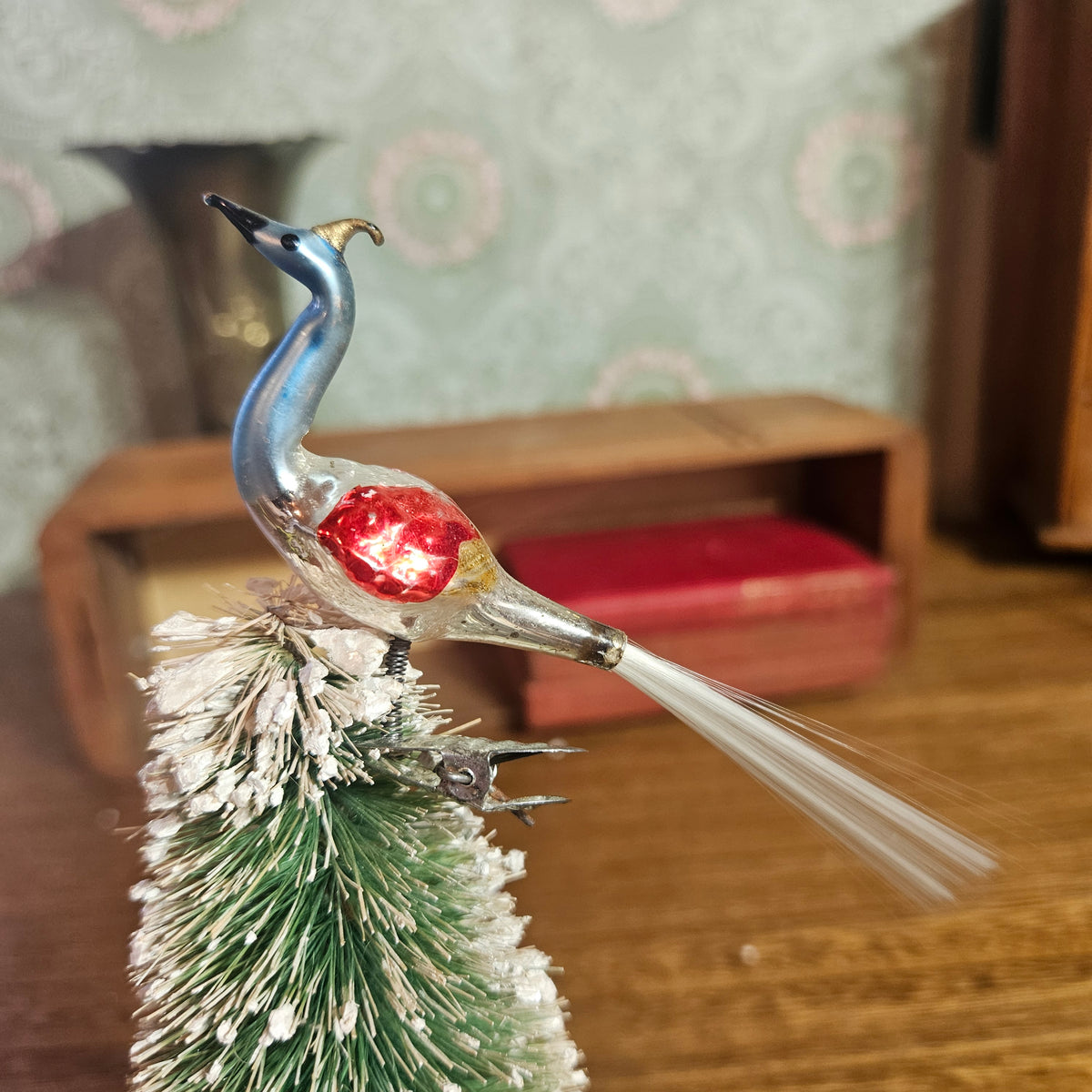A vintage-style decorative bird ornament with a blue and silver body, a red glittery chest, and long white tail feathers, perched on a small frosted green tree in a real indoor setting with blurred furniture and wallpaper in the background.