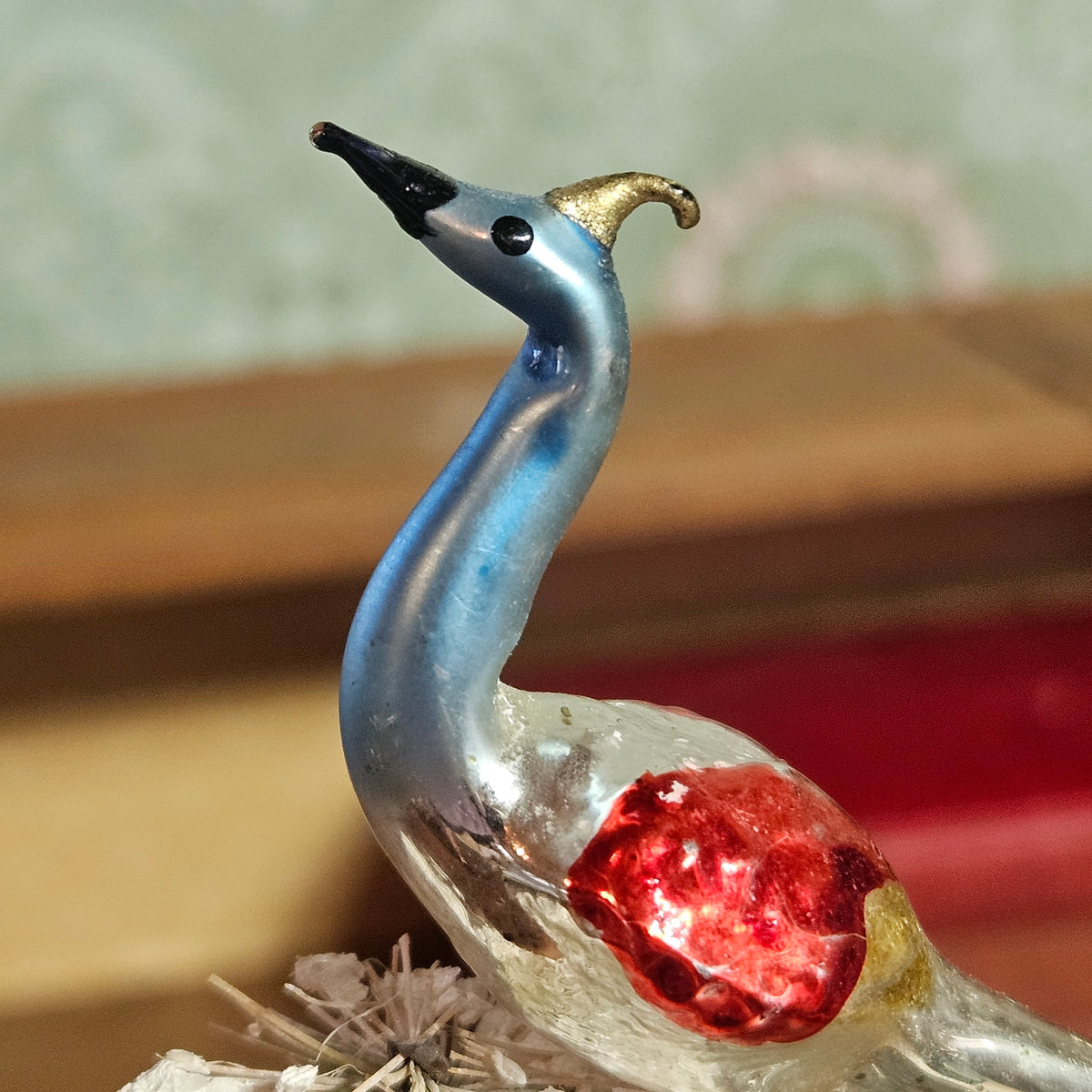 A decorative glass bird figurine with a blue neck, clear body, red glitter chest, and gold crest, photographed close up against a softly blurred indoor background.