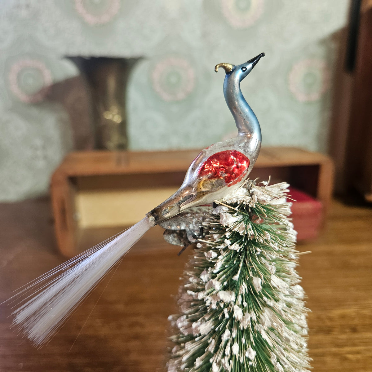 A vintage-style decorative bird ornament with a metallic blue neck, silver body, and a red glitter chest, perched on top of a small frosted green and white Christmas tree branch.