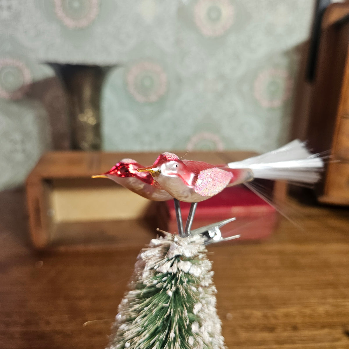A vintage-style bird ornament with a glittery pink and red body, yellow beak, and white tail feathers, perched on top of a small frosted green and white Christmas tree branch, photographed on a wooden surface with a blurred patterned wallpaper background.