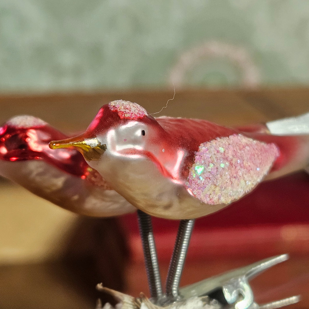 A close-up of a decorative bird ornament with a glossy red and white body, glittery pink wing, and metallic gold beak, mounted on thin metal stands.
