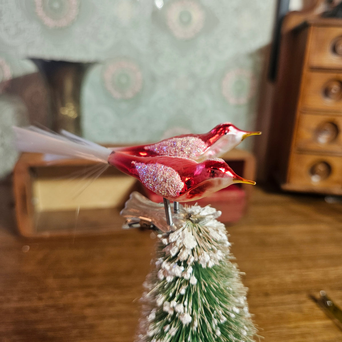 A pair of red glittery bird ornaments with white tails and yellow beaks, perched on top of a small frosted green Christmas tree, set against a warm, blurred vintage background.