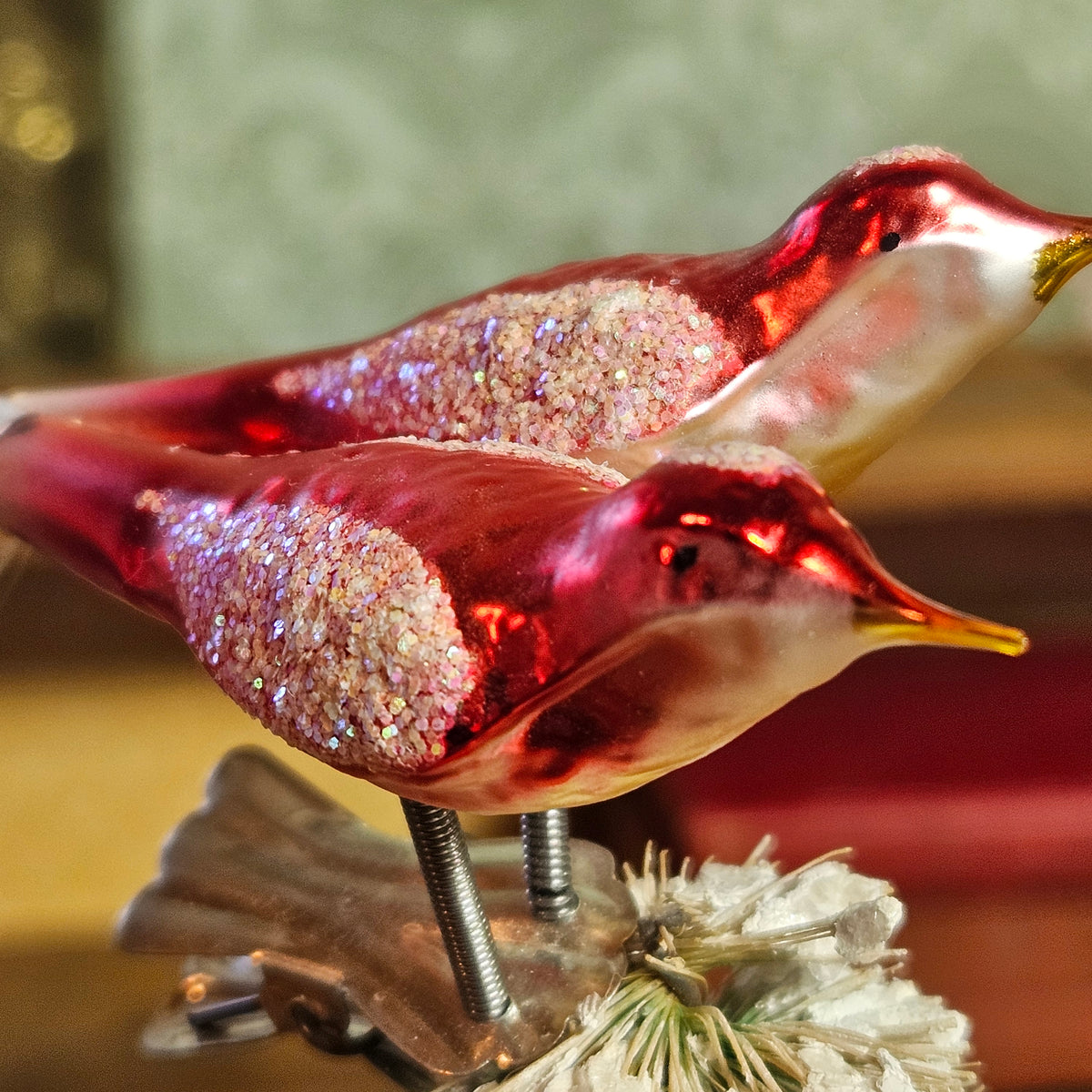 A pair of decorative bird ornaments with metallic red bodies, glittery pink wings, and yellow beaks, mounted on a stand with a dried flower.