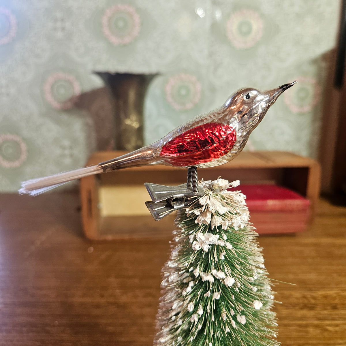 A vintage-style metal bird ornament with a shiny red chest and silver body, perched on top of a small green artificial Christmas tree with white faux snow.