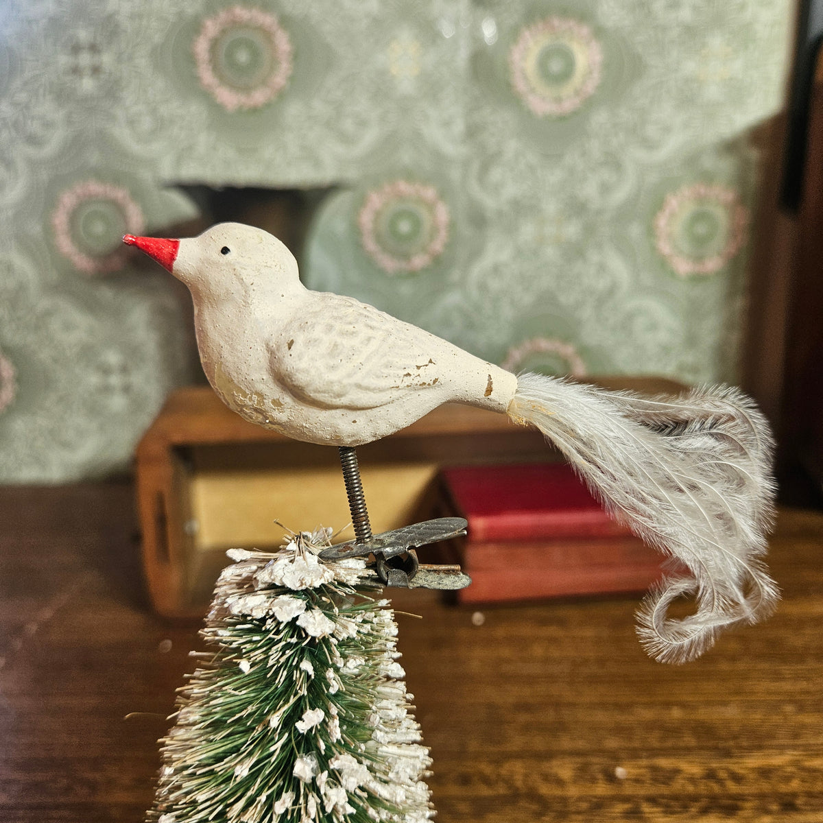 A vintage-style white bird ornament with a red beak perched on top of a small frosted Christmas tree, set on a wooden surface with patterned wallpaper and books in the background.