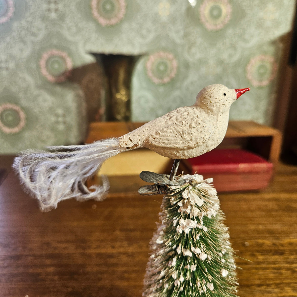 A vintage-style cream-colored bird ornament with a red beak and a fluffy white tail, perched on top of a small green and white frosted Christmas tree branch, set against a warm, softly blurred background.