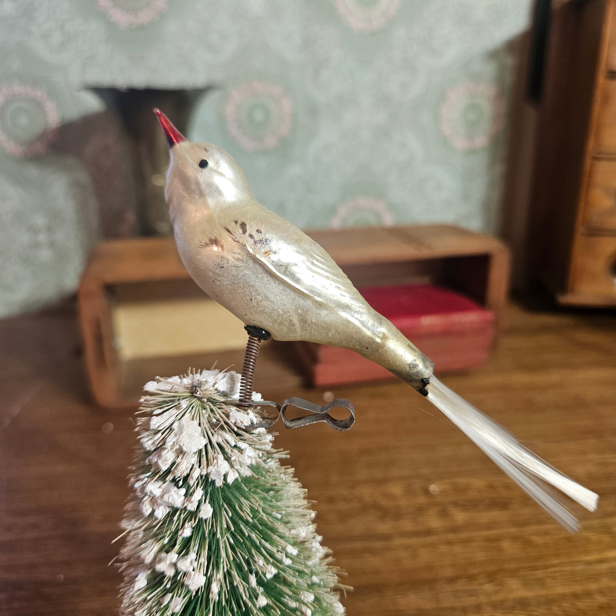A vintage metallic gold bird ornament with a red beak perched on top of a small green and white frosted Christmas tree, set against a warm, blurred background of wooden furniture and patterned wallpaper.