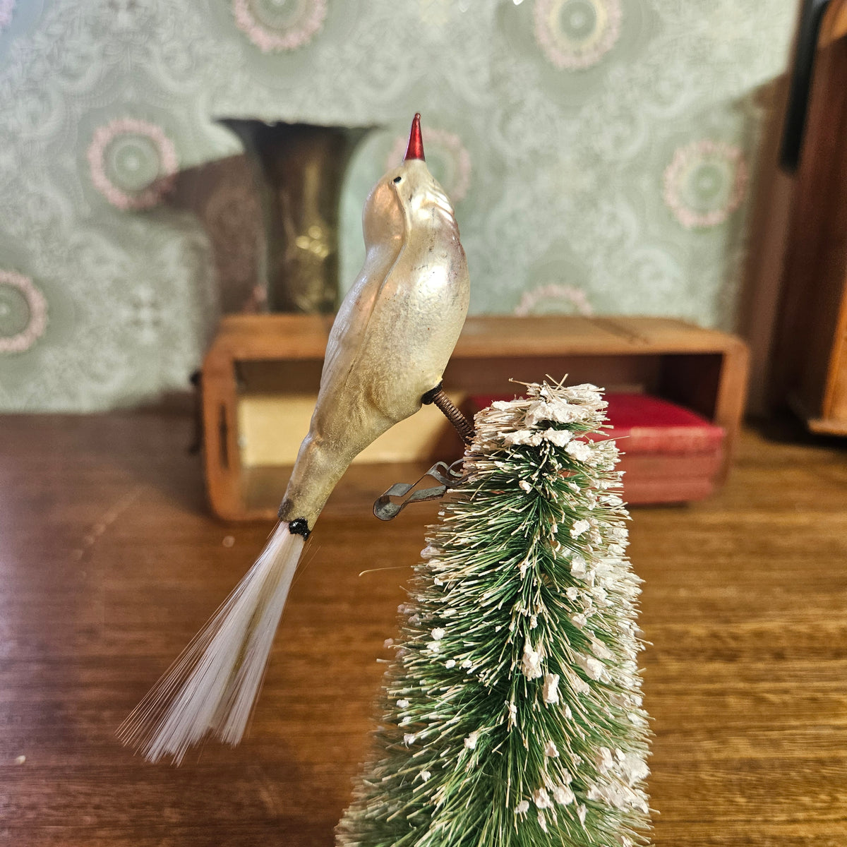 A vintage-style bird ornament with a metallic gold body, red beak, and white tail feathers, mounted on a small frosted green Christmas tree. The background shows a wooden table and a patterned wallpapered wall.