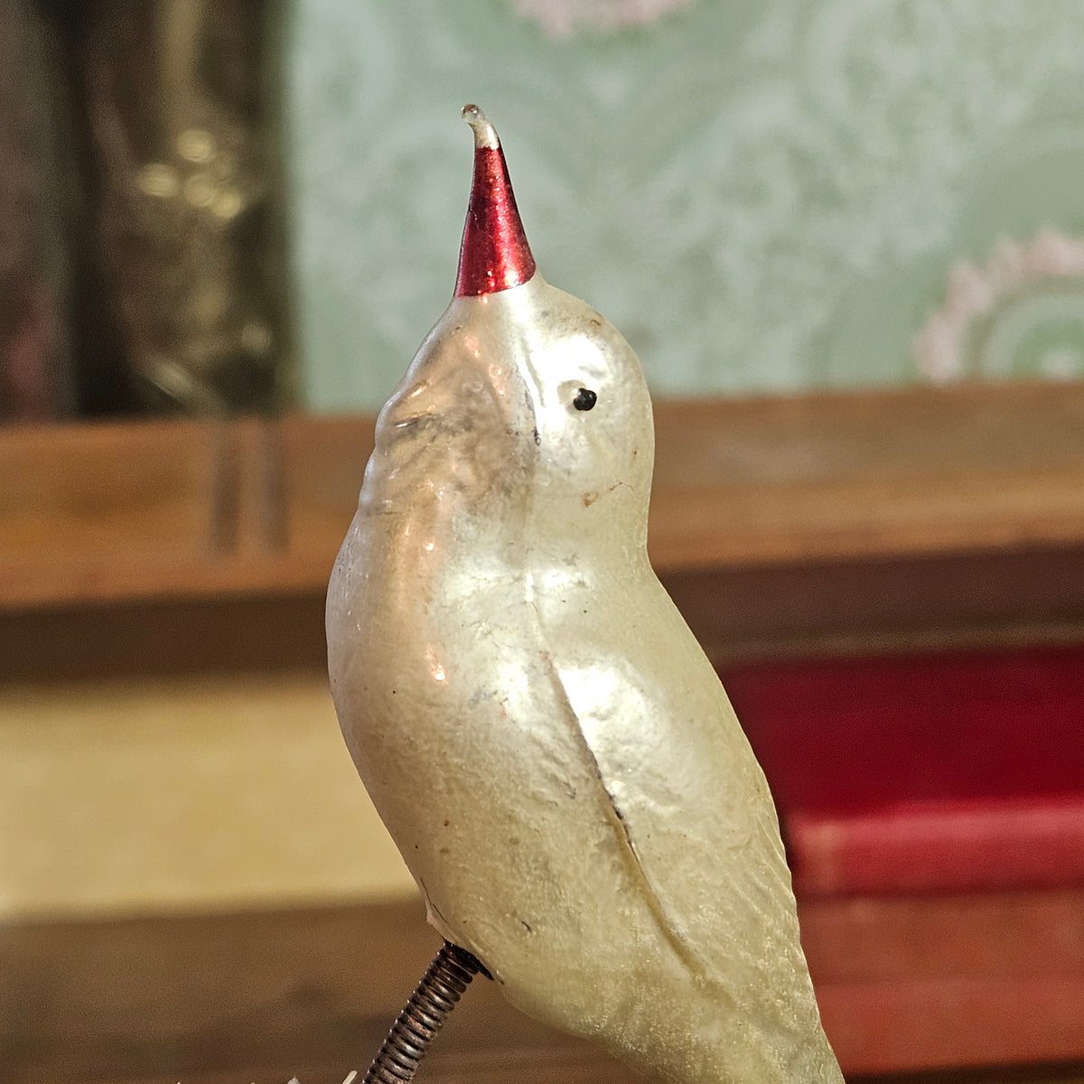 A vintage silver bird figurine with a red conical beak, perched on a metal spring against a blurred background.