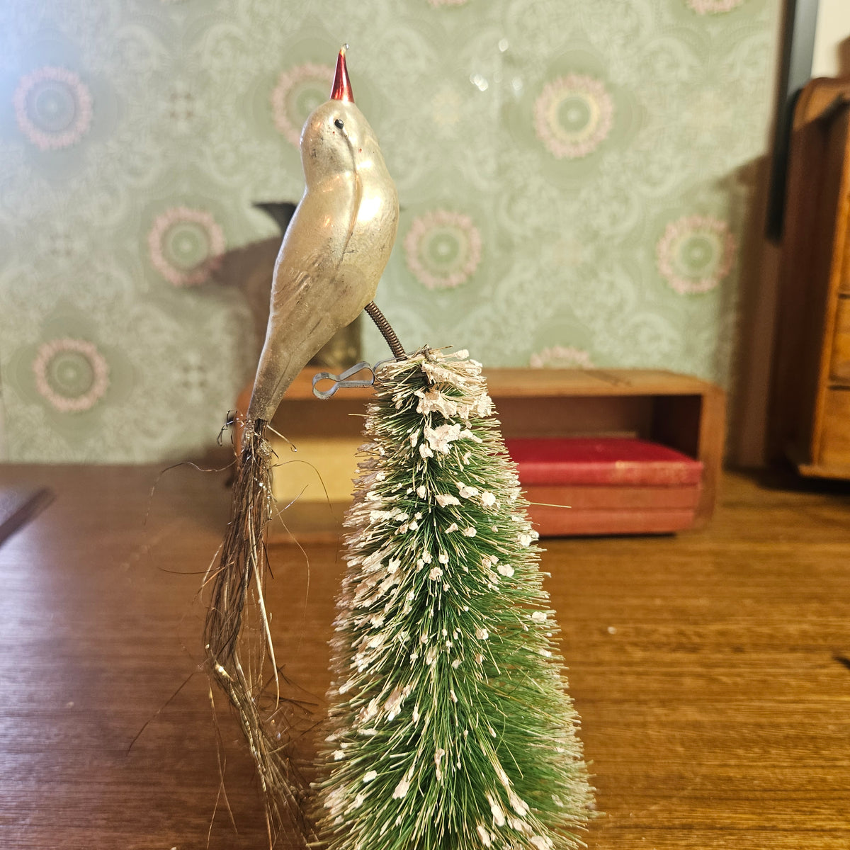 A vintage-style metal bird ornament perched on top of a small green bottlebrush Christmas tree with white tinsel snow, displayed on a wooden table against a patterned wallpaper background.