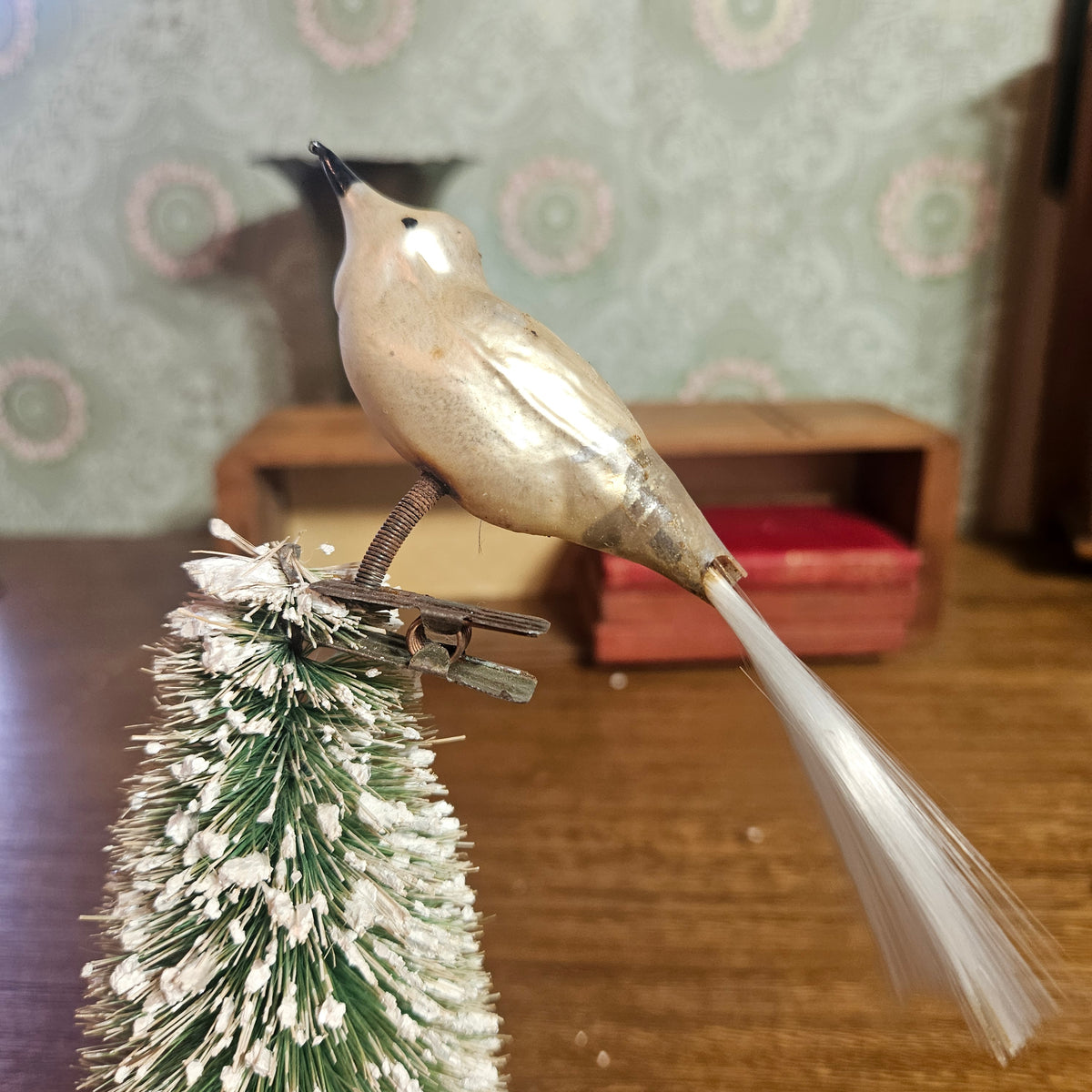A vintage metal bird ornament with a long white tail, perched on top of a small green and white frosted Christmas tree branch, set against a blurred background of patterned wallpaper and wooden furniture.