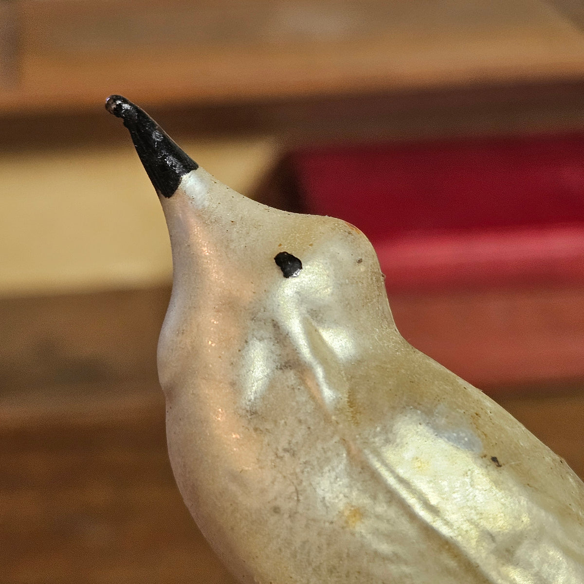 A close-up of a ceramic bird figurine with a glossy cream-white body, a small black beak, and a tiny black eye detail, set against a blurred background of wood and red tones.