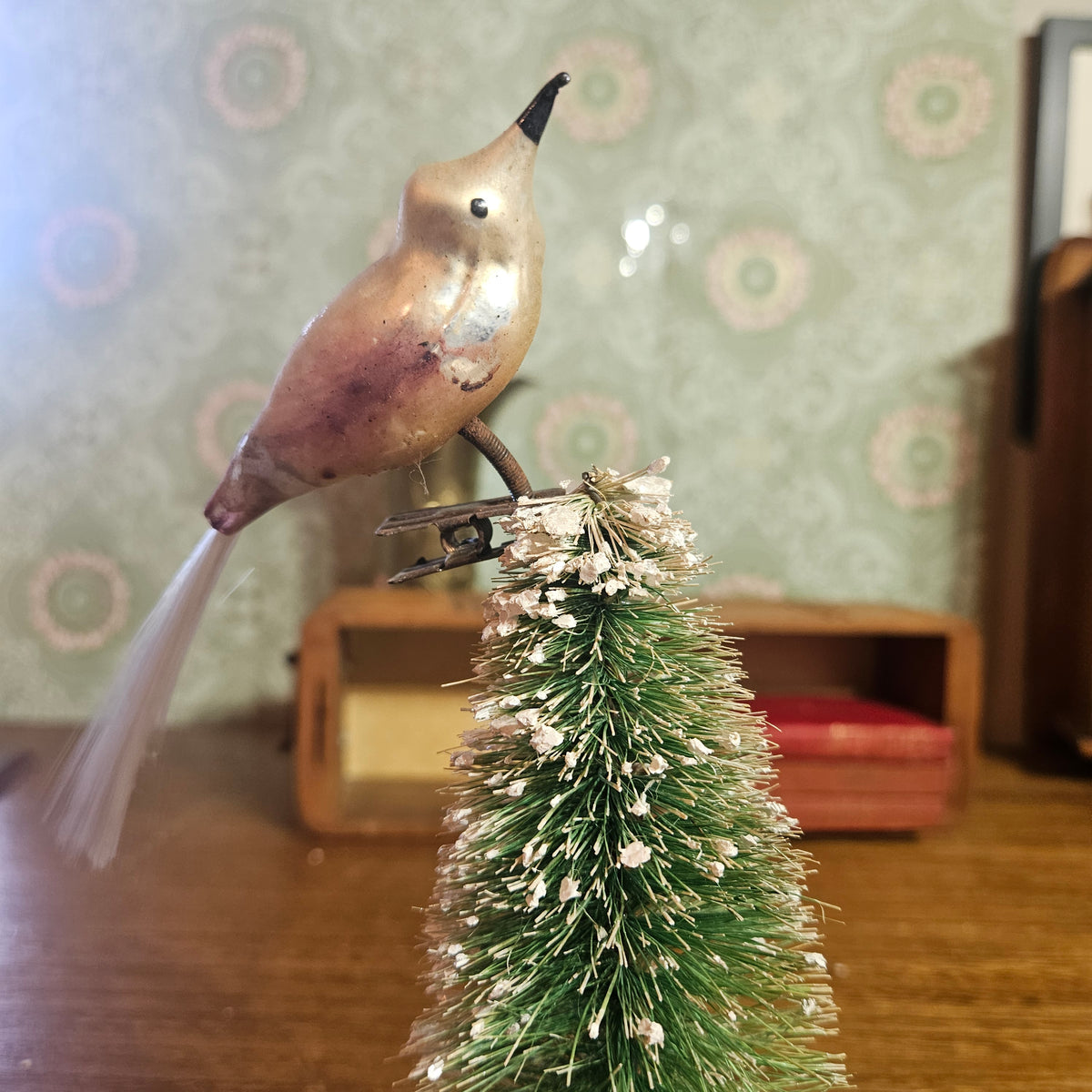 A vintage-style metal bird ornament with a metallic bronze finish and a black beak, perched on top of a small green bottlebrush Christmas tree with white snow-like accents, set against a patterned wallpaper background.