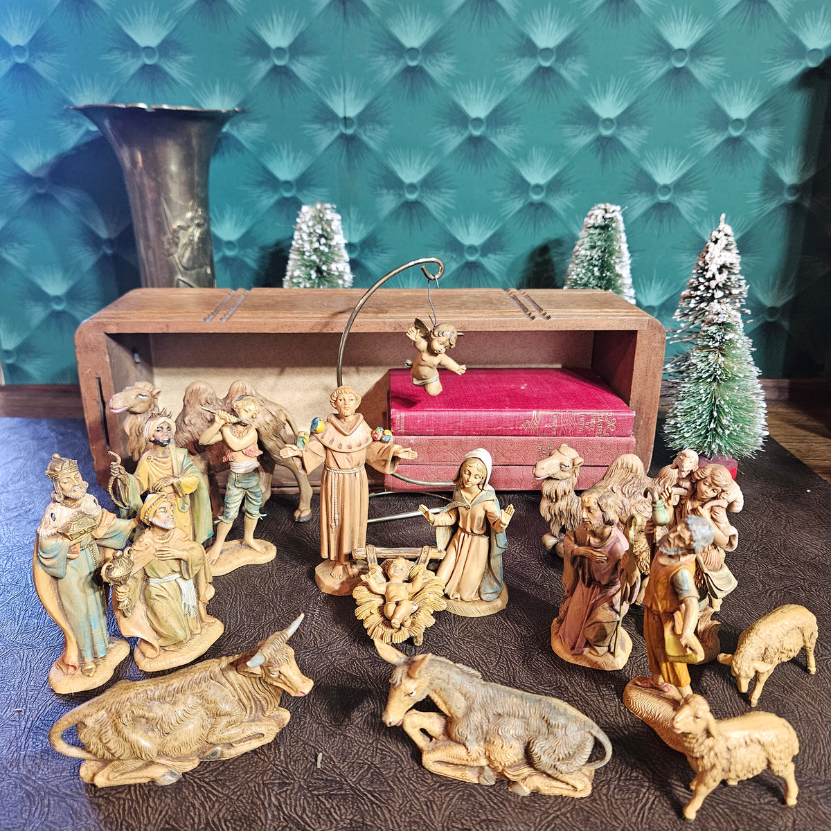 A vintage wooden nativity set with multiple hand-painted figurines, including Mary, Joseph, baby Jesus, the Three Wise Men, shepherds, and animals, arranged in front of a small wooden stable with a red cushion and miniature trees.