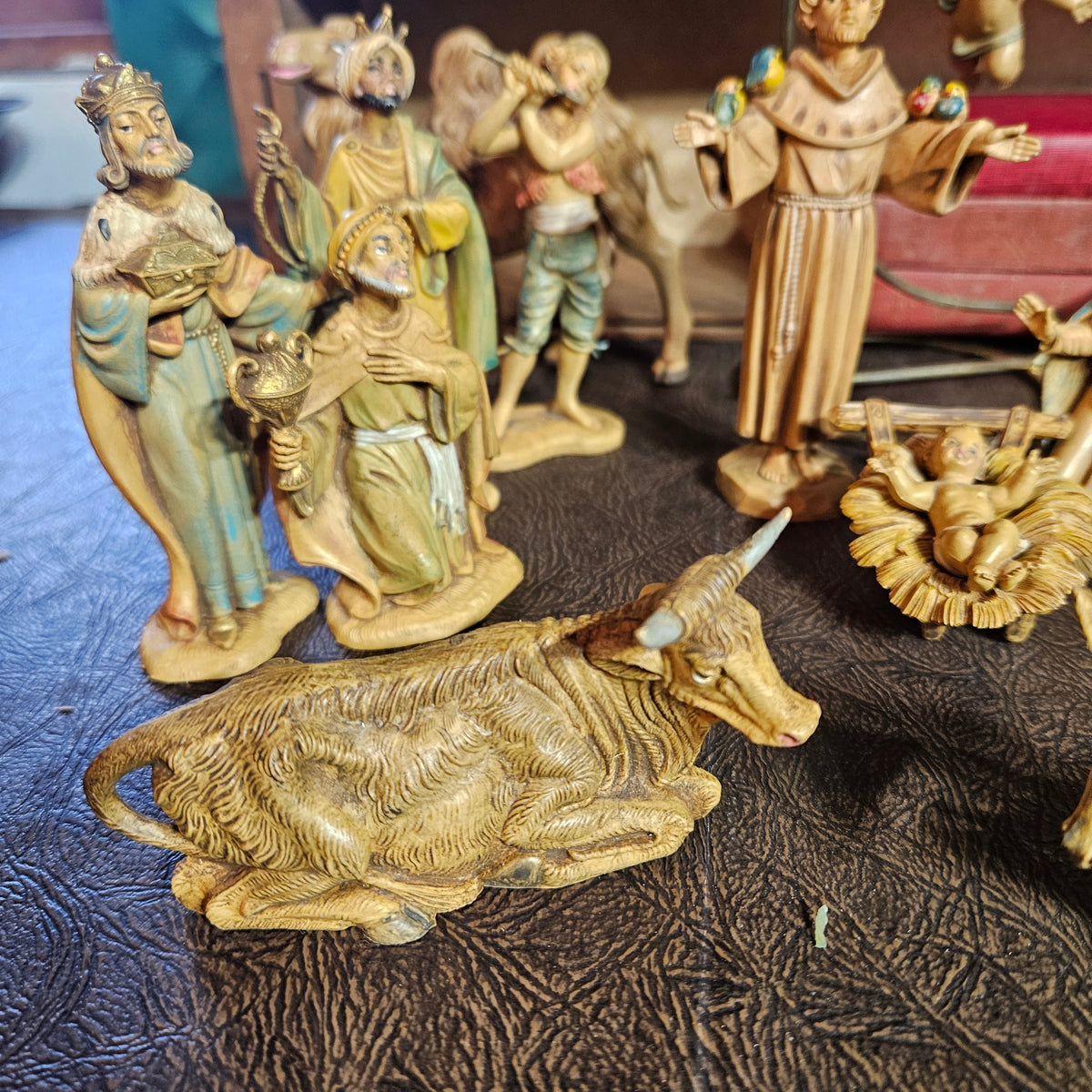 A set of hand-carved wooden nativity figurines displayed on a dark textured surface. The set includes a卧ing cow, a baby in a manger, and several wise men and saints with painted details.