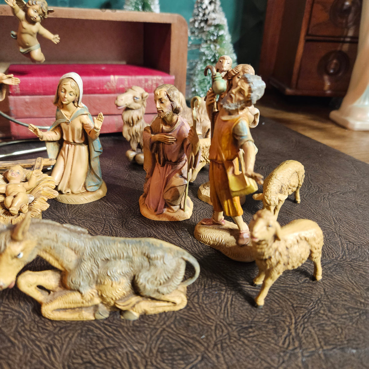 A set of hand-painted wooden nativity figurines arranged on a dark textured surface, including Mary, Joseph, baby Jesus, an angel, and animals like sheep and a donkey, with a wooden shelf and Christmas tree in the background.