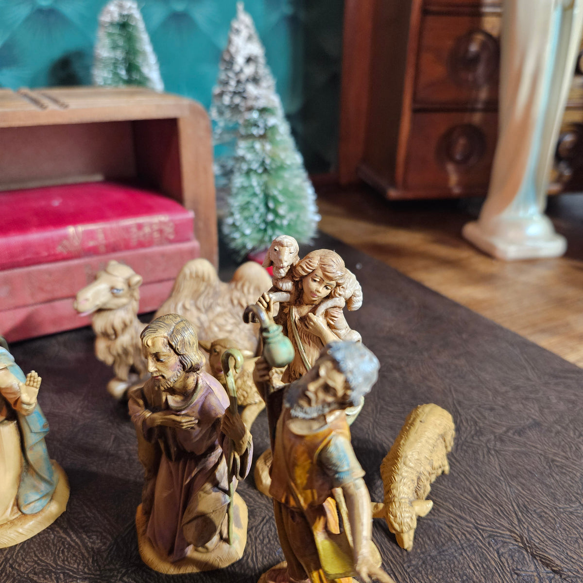 A set of hand-carved wooden nativity figurines arranged on a dark textured surface, with a vintage wooden cabinet, red box, and small artificial trees in the background.