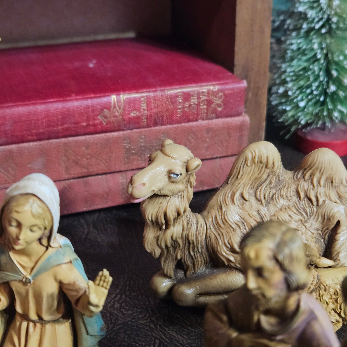 A close-up of a rustic nativity scene featuring a carved camel figurine, a child with a blue cloak, and another figure, set against a background of vintage red books and a small green tree.