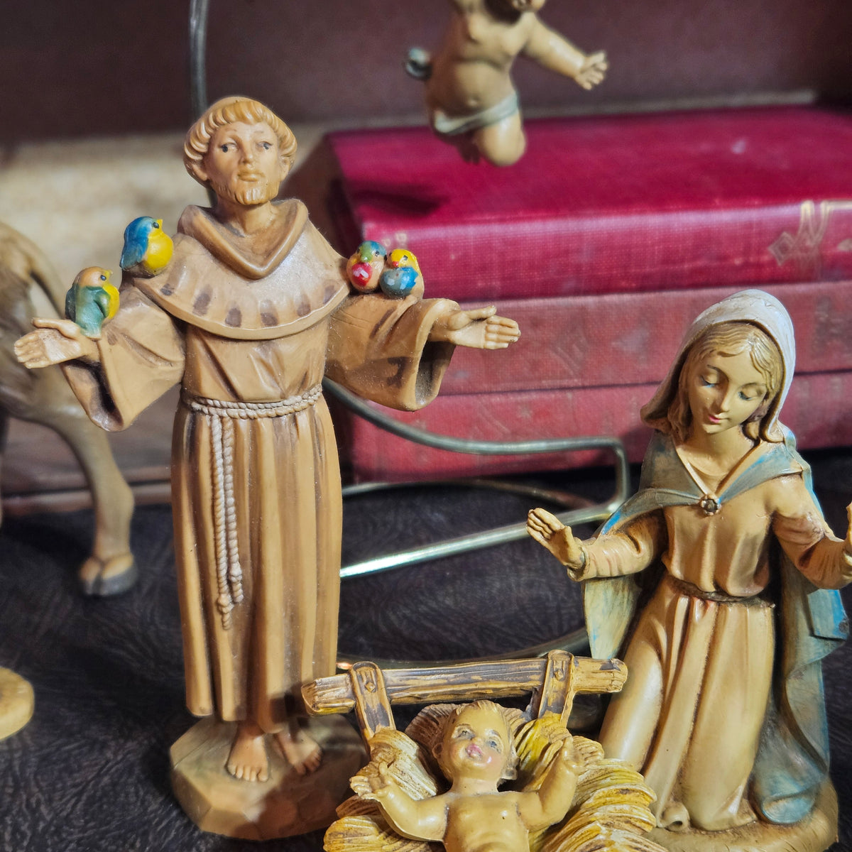 A wooden nativity scene figurine set featuring Saint Joseph, Mary, and baby Jesus in a stable, with small colorful birds on Saint Joseph's shoulders, displayed on a dark surface with a red box in the background.