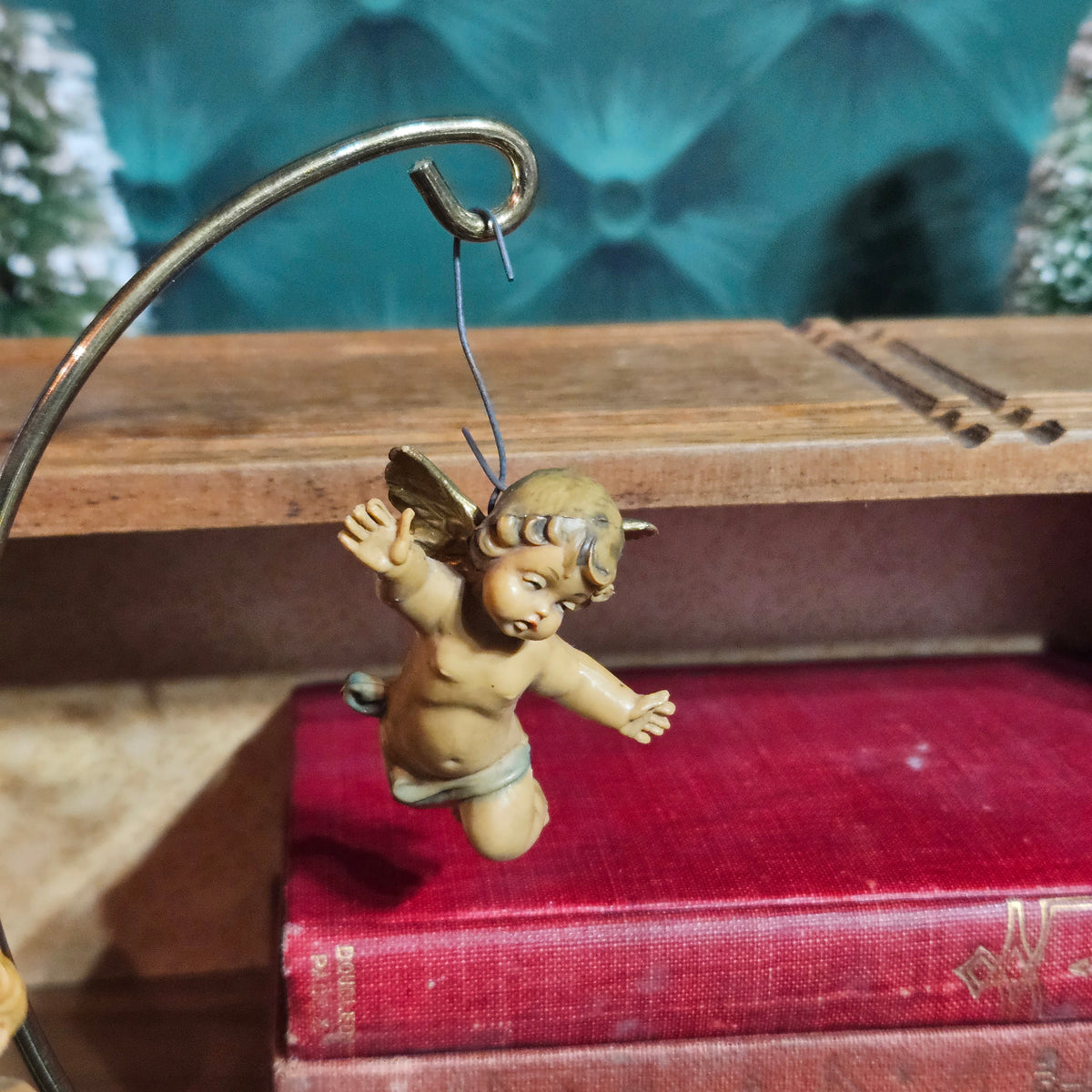 A small cherub angel hanging ornament in a vintage tan and brown finish, suspended from a metal wire hanger on a wooden shelf above a red book.