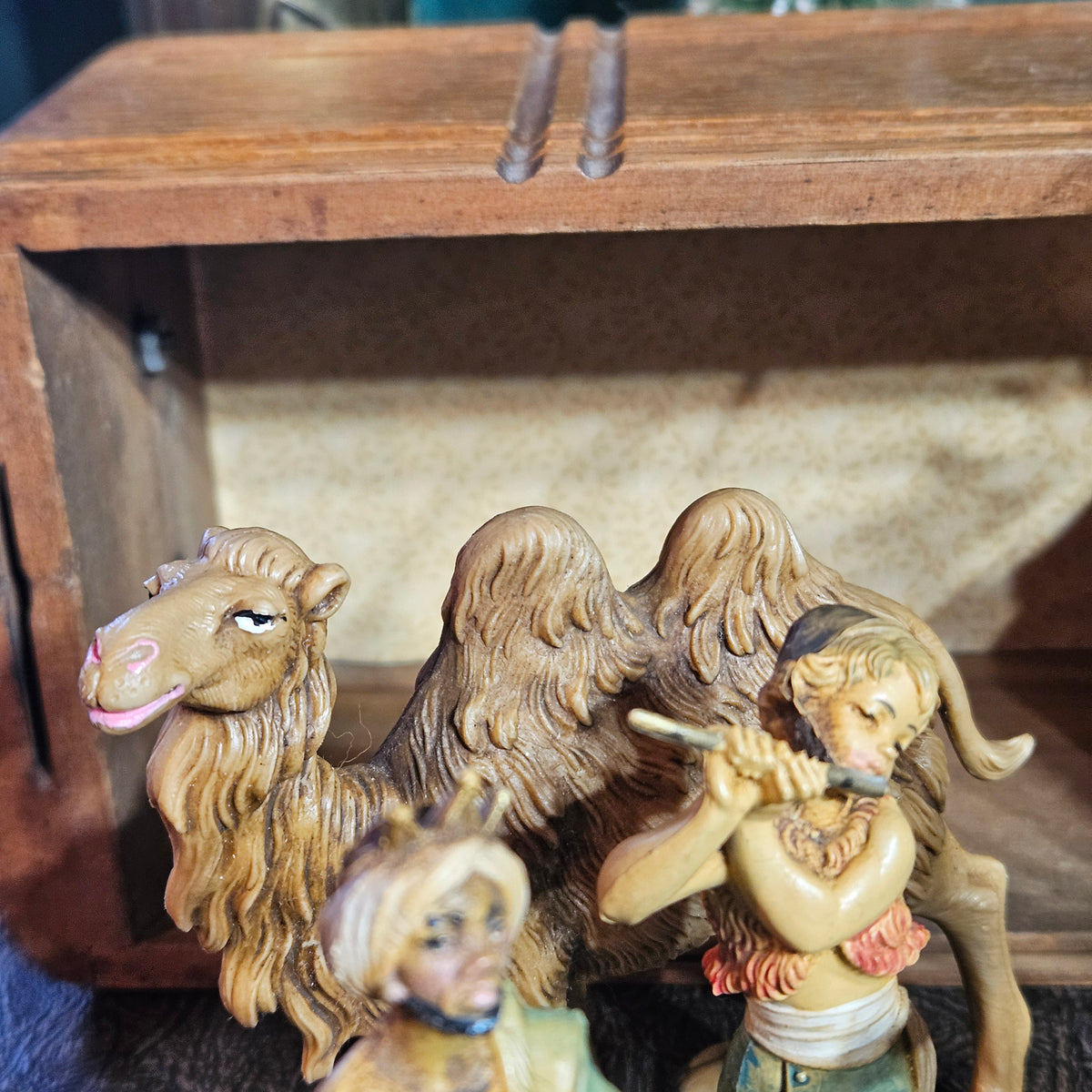 A small decorative figurine set on a wooden shelf, featuring a camel, a robed figure holding a pipe, and another partially visible figure in the foreground.