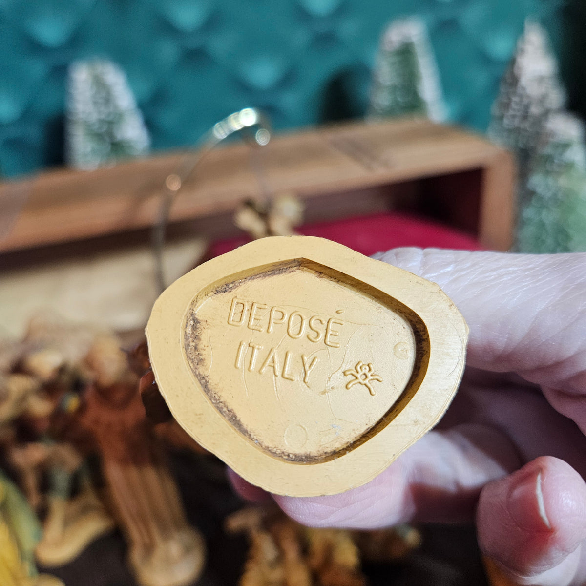 A hand holding a gold-colored metal coin blank with 'DEPOSED ITALY' and a small star engraved on the reverse side, with a blurred background showing miniature decorative figures and a teal textured backdrop.