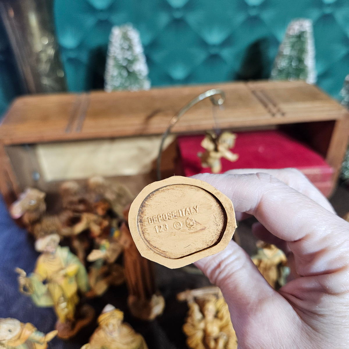 A hand holds a small carved wooden ornament or figurine piece showing the bottom end, which is stamped with 'Depose Italy 123'. The background shows a wooden display case with red velvet lining and other carved wooden figures.