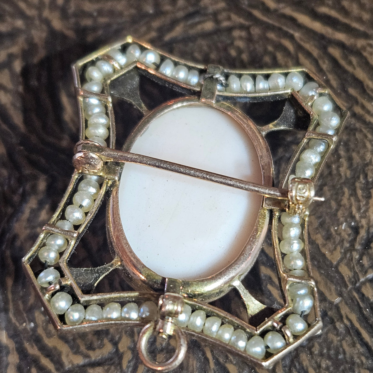 An antique-style gold-toned brooch featuring a central oval white enamel inlay, surrounded by a border of small white pearls and geometric cutout detailing, photographed on a dark wood background.