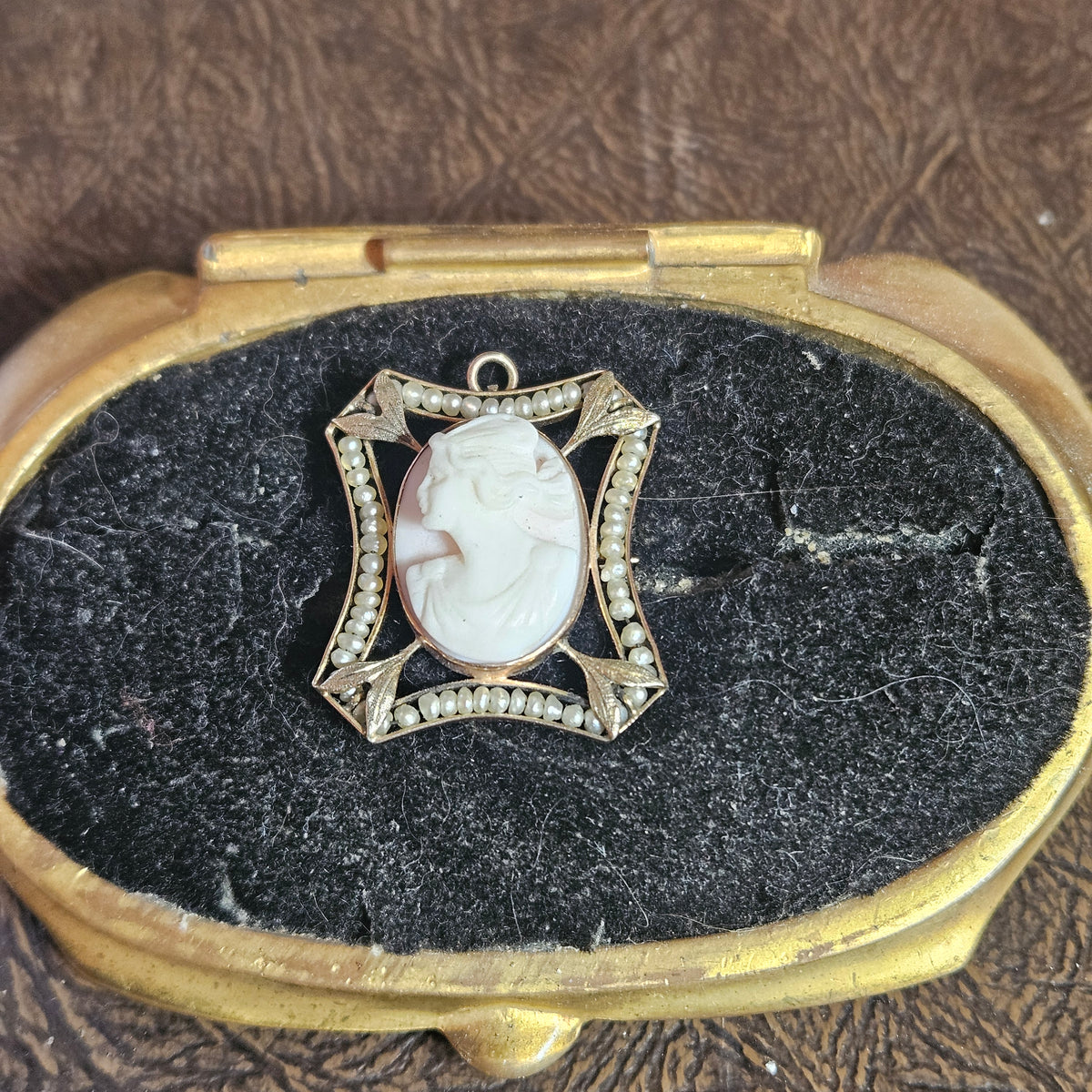 An antique-style oval pendant with a carved white cameo face in a gold-toned filigree frame, displayed on a black velvet-lined gold case over a brown textured surface.