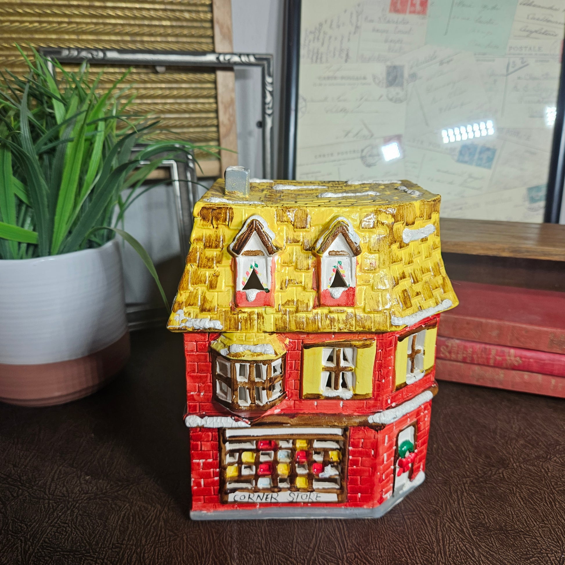 Decorative house-shaped container on a surface with a plant and books in the background