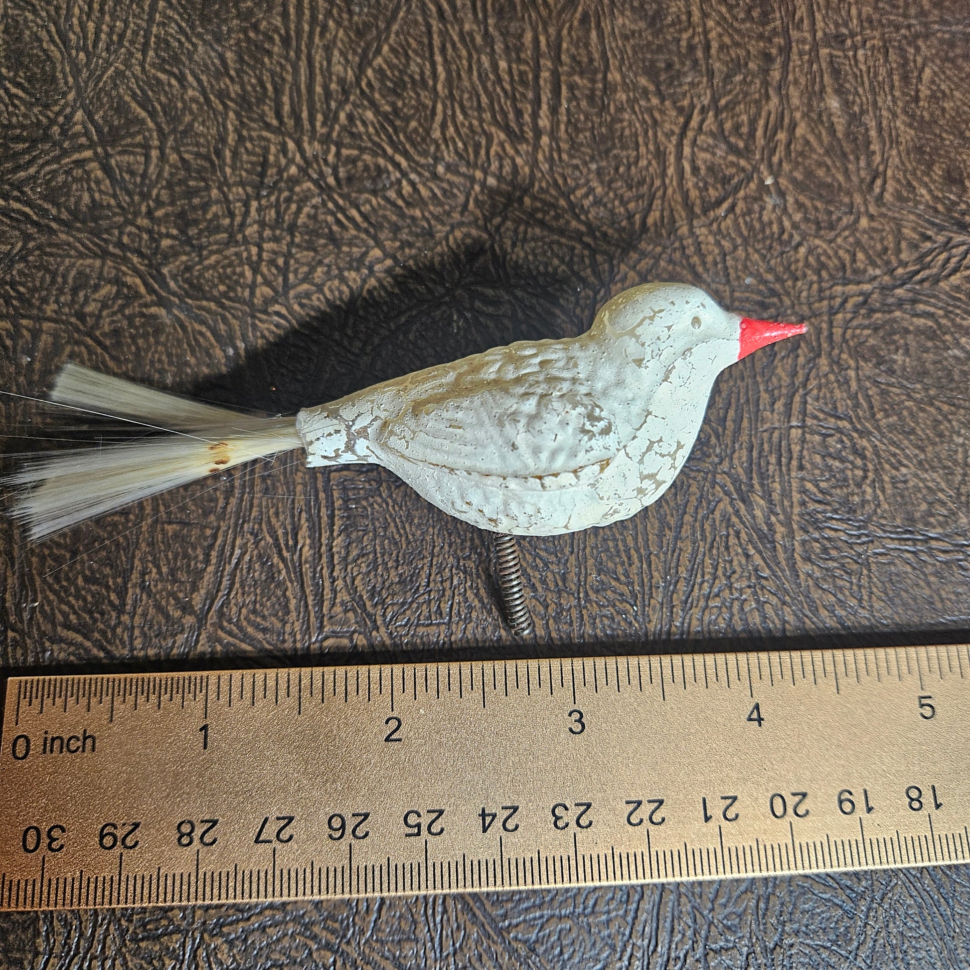 Decorative bird ornament with a red beak next to a ruler on a textured surface